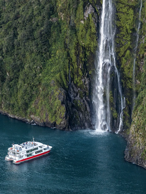 Cruise boat near waterfall in Milford Sound, New Zealand, surrounded by lush cliffs.