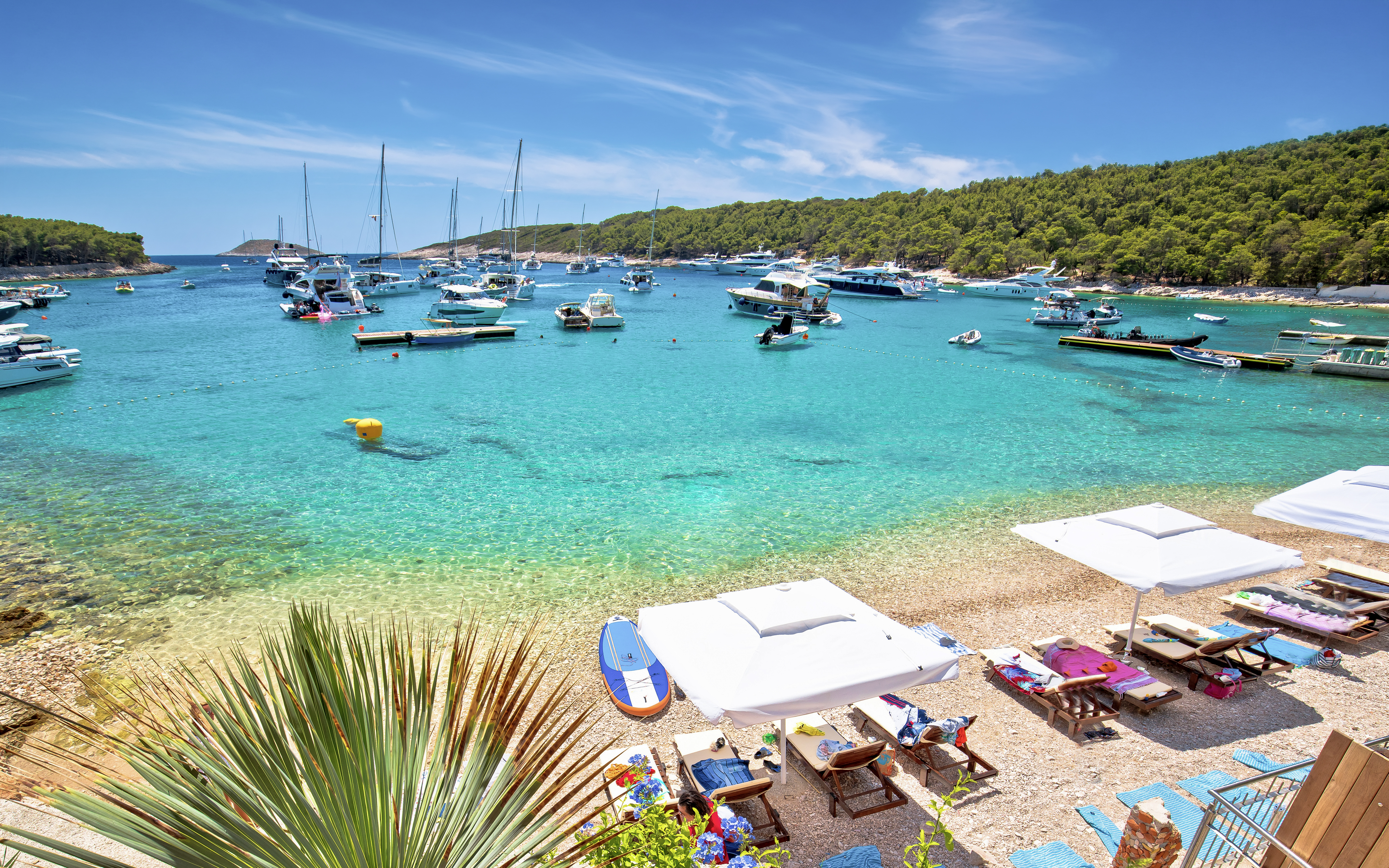 Yachts anchored at Palmizana turquoise beach, Pakleni Otoci islands, with sun loungers and umbrellas.
