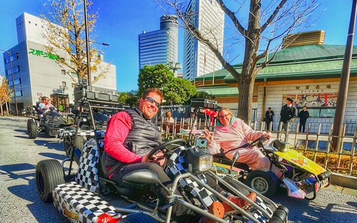 Two people riding go-karts on a Tokyo street with city buildings in the background.