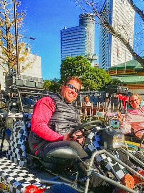 Two people riding go-karts on a Tokyo street with city buildings in the background.