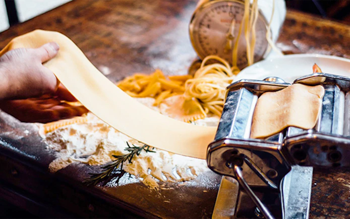 Pasta being rolled through a machine on a wooden table with flour and rosemary.