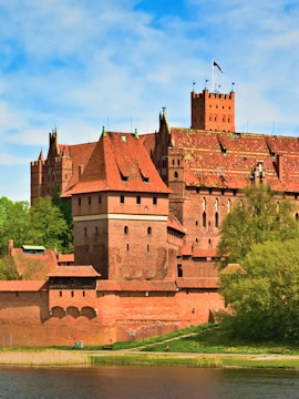 Malbork Castle's red brick architecture with towers and a flag, viewed from the river.