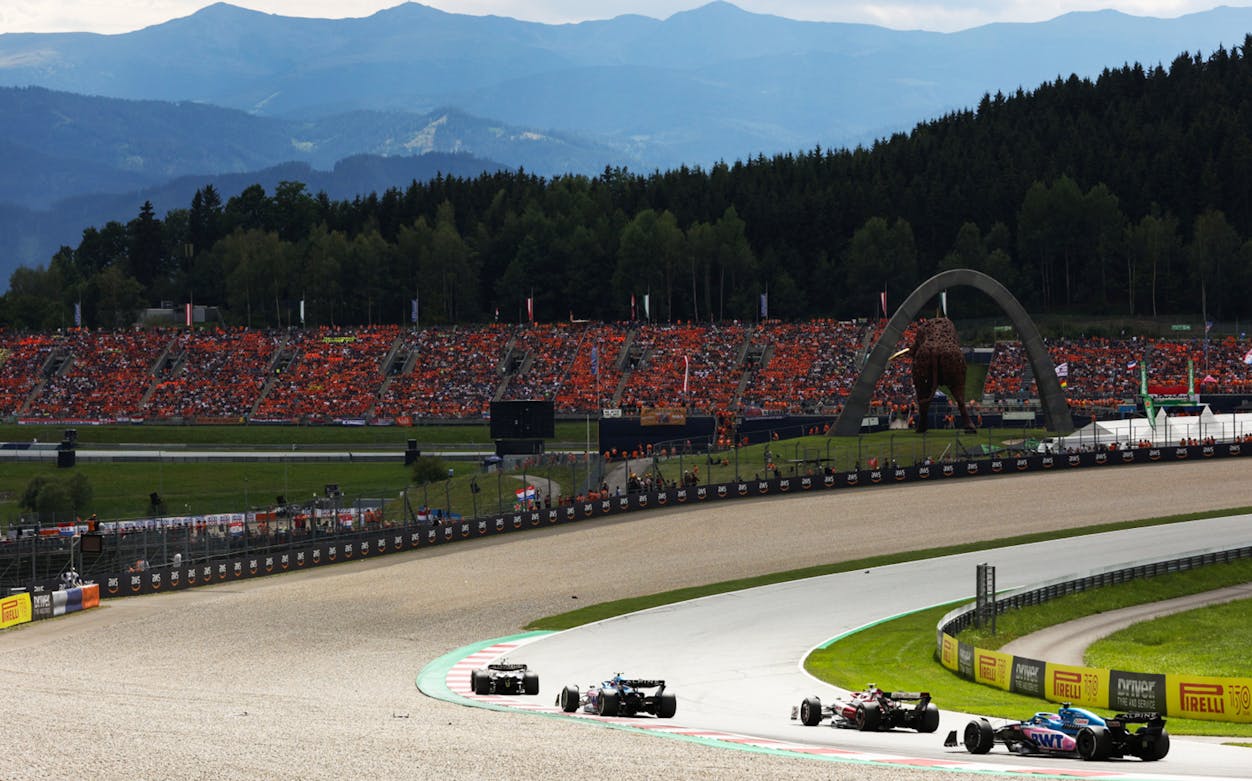 Formula 1 cars racing at the Austrian Grand Prix with a packed grandstand and mountainous backdrop.