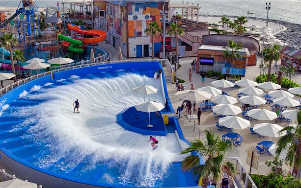 Surfers on wave simulator at Laguna Waterpark, Dubai, with colorful slides and seating area.