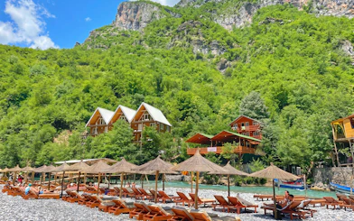 Sun loungers and wooden cabins by Shala River, surrounded by lush green hills, Komani Lake day trip.