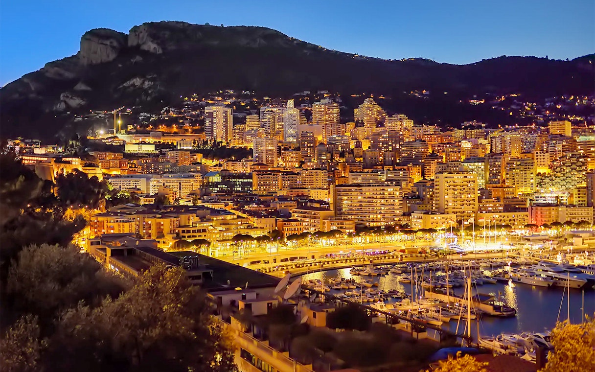 Monaco skyline at night with illuminated buildings and marina, viewed from Nice.