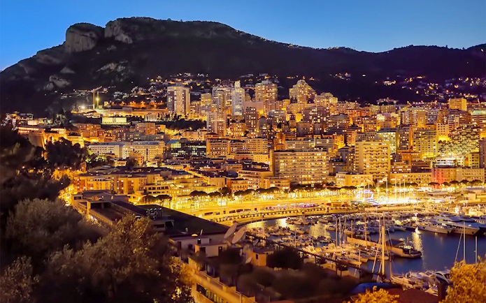Monaco skyline at night with illuminated buildings and marina, viewed from Nice.