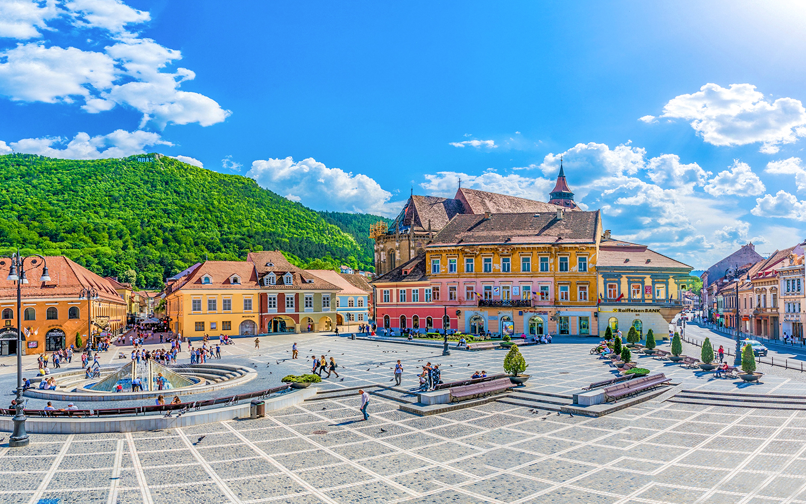 Brasov old town square with colorful buildings and people, surrounded by green hills.
