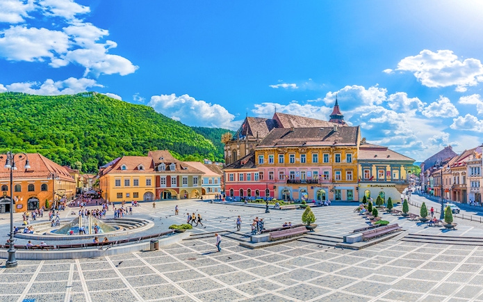 Brasov old town square with colorful buildings and people, surrounded by green hills.