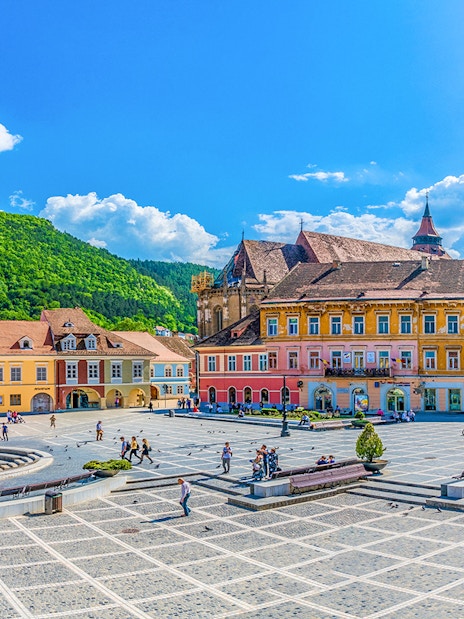 Brasov old town square with colorful buildings and people, surrounded by green hills.