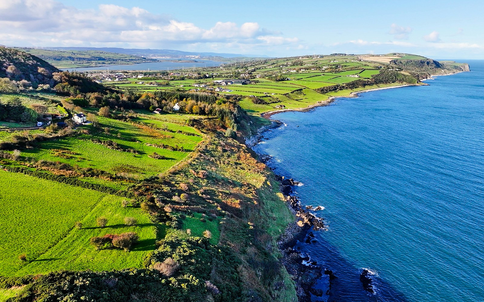 Aerial view of Glens of Antrim coastal road with lush green fields and blue sea.