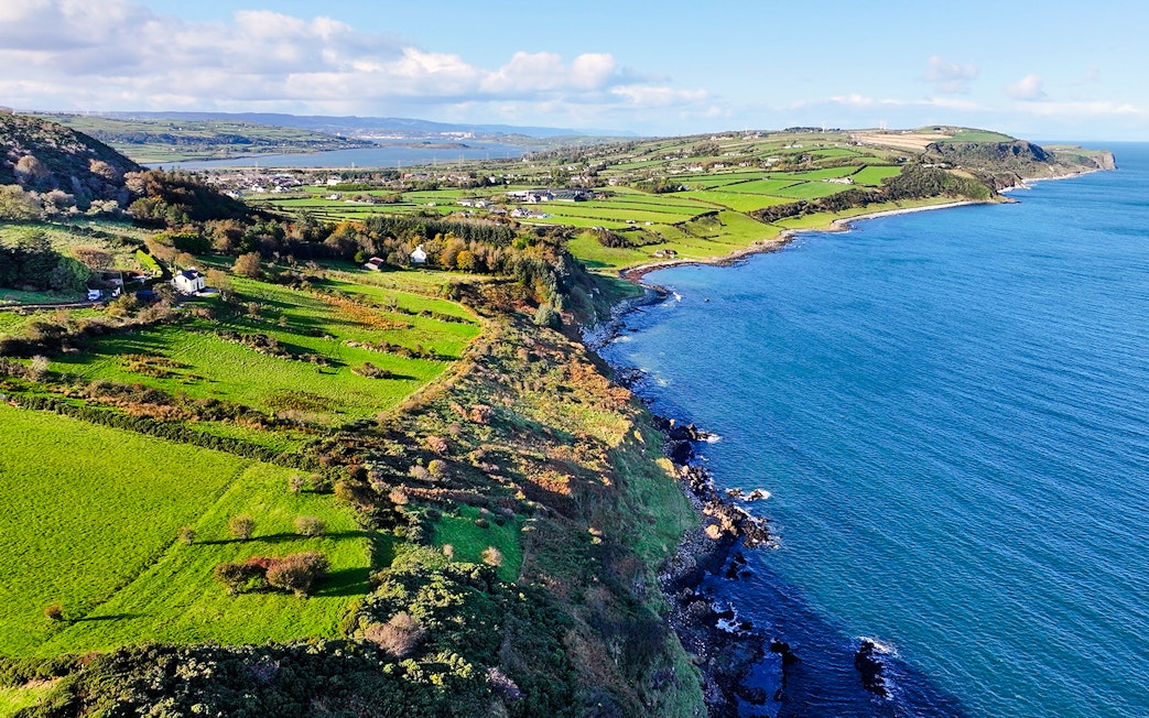 Aerial view of Glens of Antrim coastal road with lush green fields and blue sea.