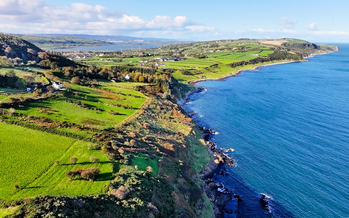 Aerial view of Glens of Antrim coastal road with lush green fields and blue sea.
