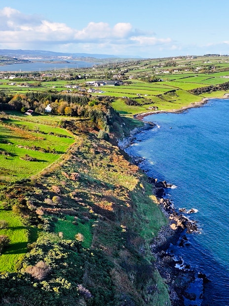Aerial view of Glens of Antrim coastal road with lush green fields and blue sea.
