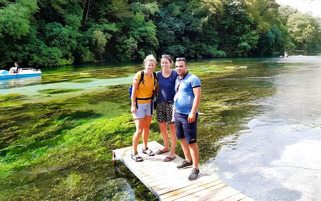 Guests standing on a wooden platform at Blue Eye springs, Albania, with lush greenery and clear water.