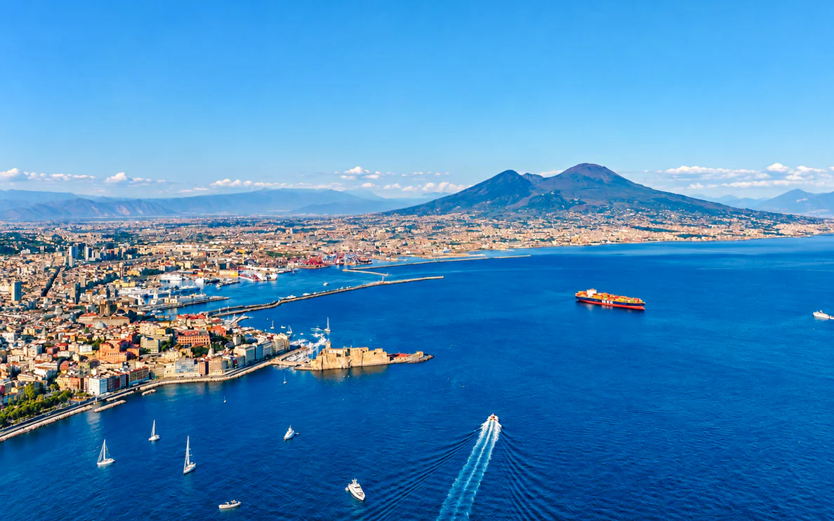 Aerial view of Naples coastline with boats heading towards Capri, featuring Mount Vesuvius in the background.