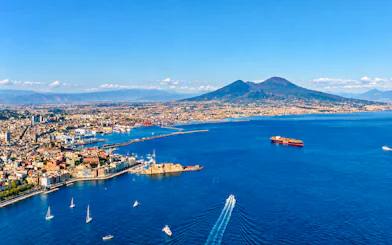 Aerial view of Naples coastline with boats heading towards Capri, featuring Mount Vesuvius in the background.