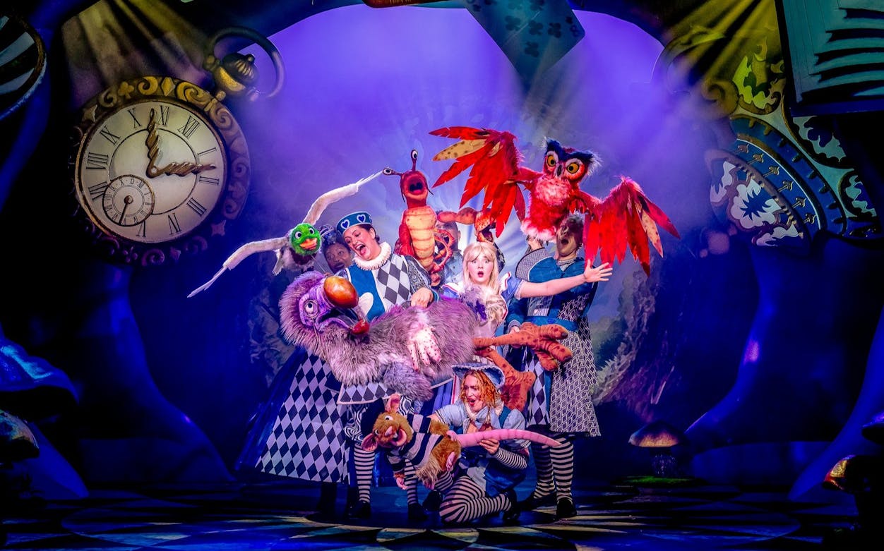Stage performance of Alice in Wonderland with colorful characters and large clock backdrop.