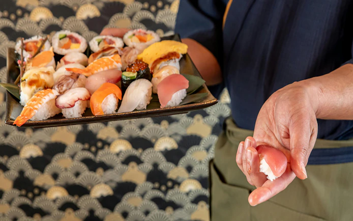 Assorted sushi platter with hand holding a piece, related to Sumo Practice Show at Ochiizumibeya.