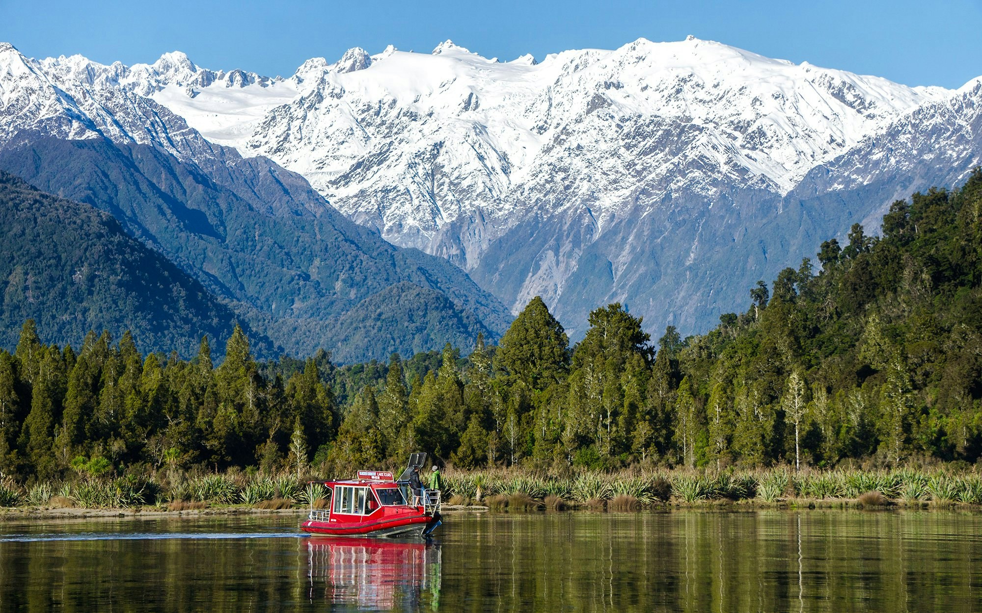 Red boat cruising on Lake Mapourika with snow-capped mountains in the background.