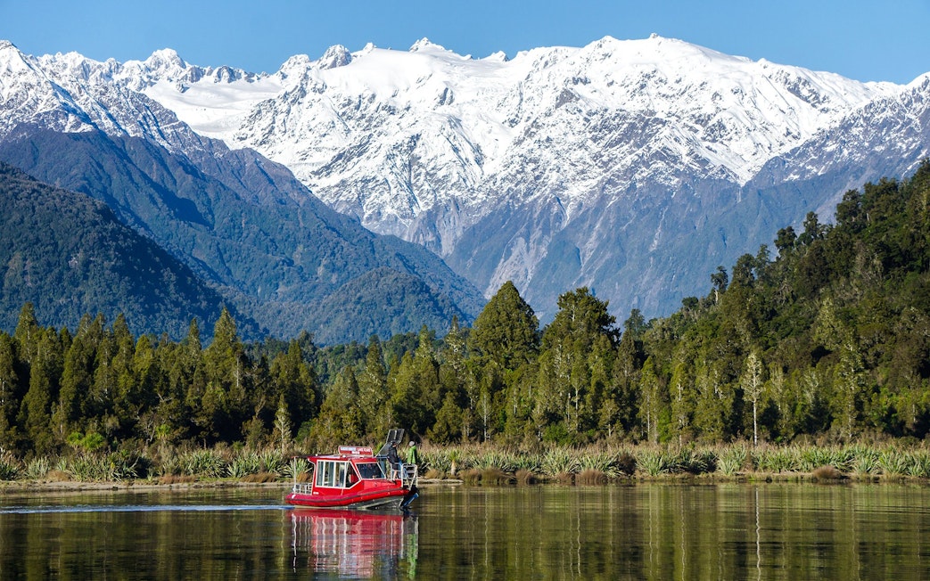 Red boat cruising on Lake Mapourika with snow-capped mountains in the background.