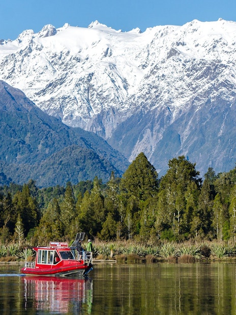 Red boat cruising on Lake Mapourika with snow-capped mountains in the background.