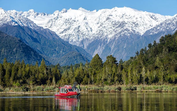 Red boat cruising on Lake Mapourika with snow-capped mountains in the background.