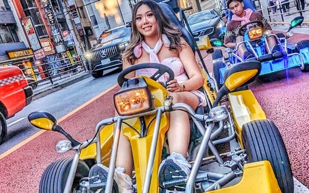 Woman in pink outfit driving a yellow go-kart on a Tokyo street.