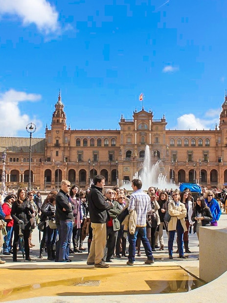 Group of tourists at Spain Square, Seville, with a guide near a fountain and historic building.