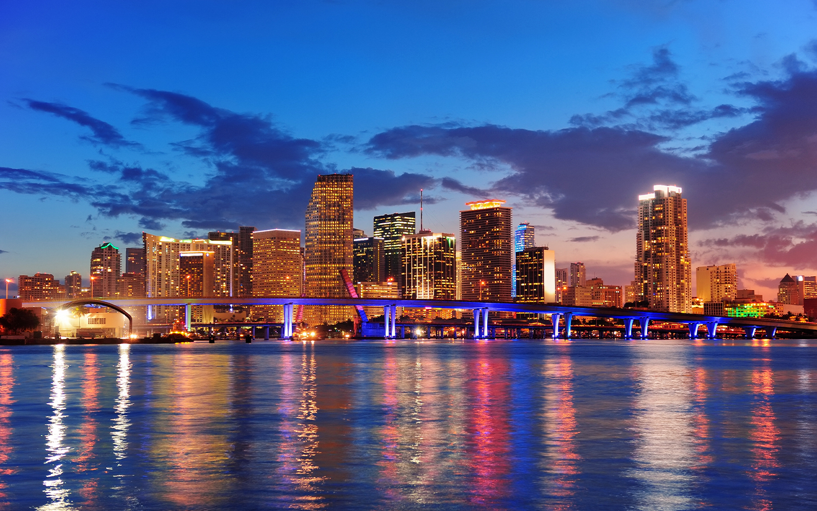 Miami city skyline at night with illuminated buildings and bridge reflecting on water.