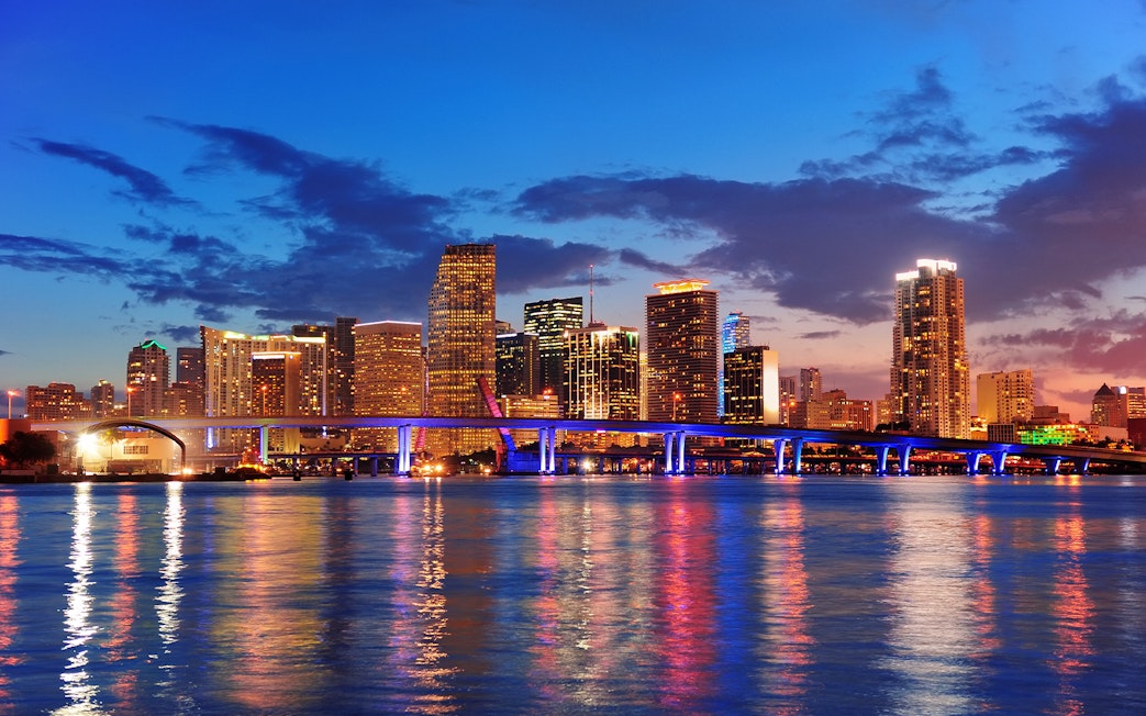 Miami city skyline at night with illuminated buildings and bridge reflecting on water.