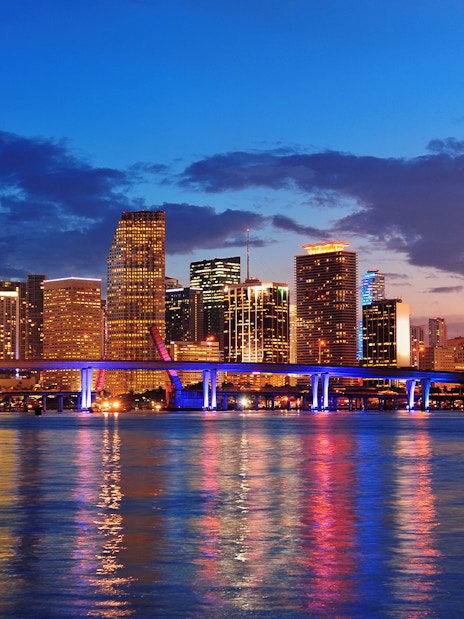 Miami city skyline at night with illuminated buildings and bridge reflecting on water.