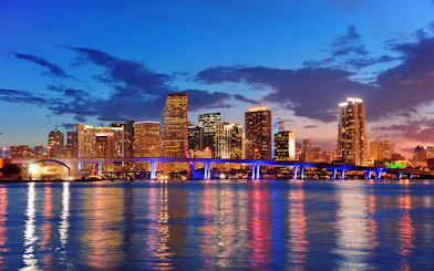 Miami skyline at dusk with illuminated bridge reflecting on water.