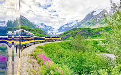 Alaska Railroad train traveling through lush green mountains with snow-capped peaks.