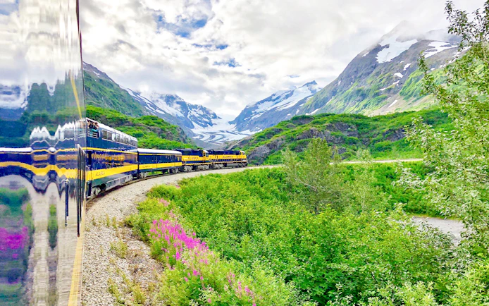 Alaska Railroad train traveling through lush green mountains with snow-capped peaks.