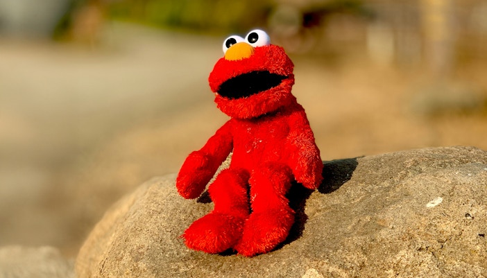 Elmo plush toy sitting on a rock outdoors.