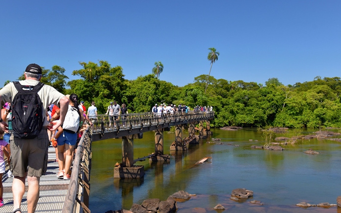 Visitors walking on a bridge towards Iguazu Falls, surrounded by lush greenery.