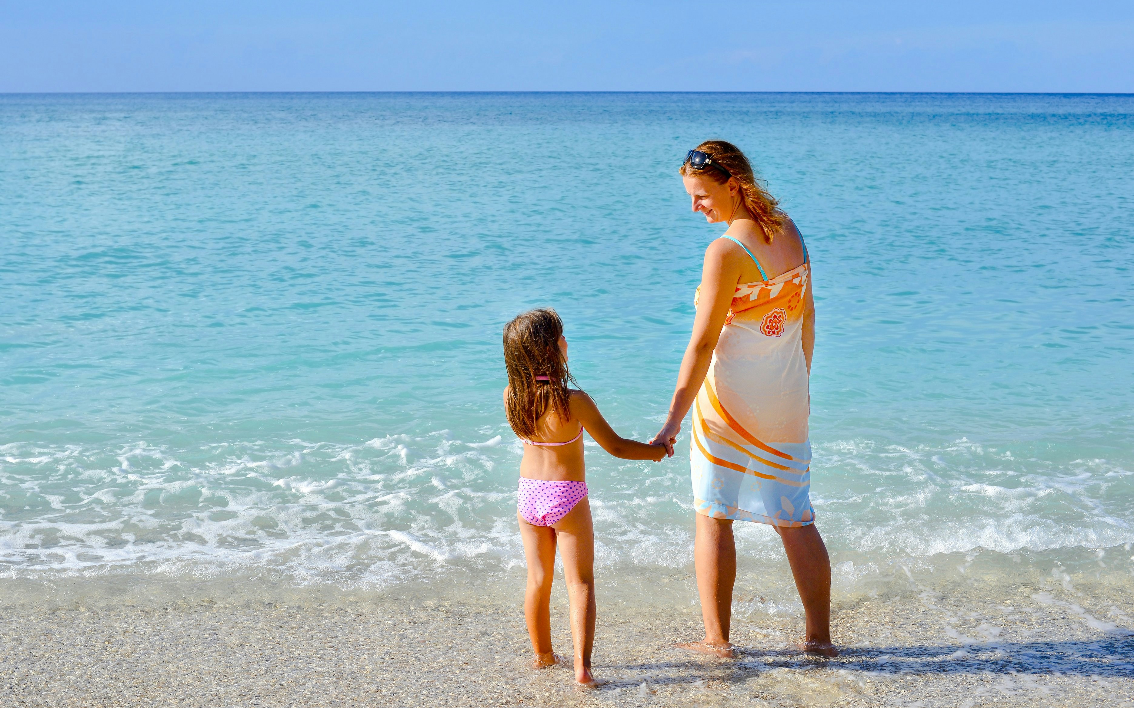 Mother and daughter holding hands at the beach, facing the ocean.