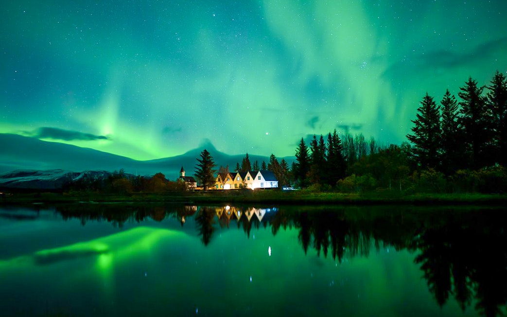Northern lights over a serene landscape with trees and a small village, seen during a Super Jeep tour.