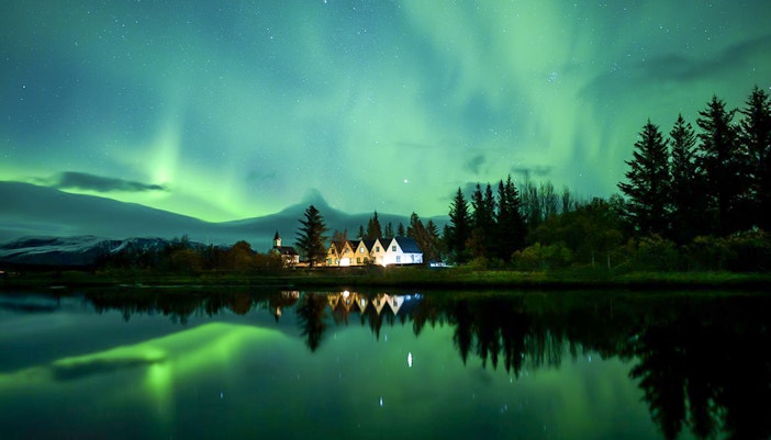 Northern lights over a serene landscape with trees and a small village, seen during a Super Jeep tour.