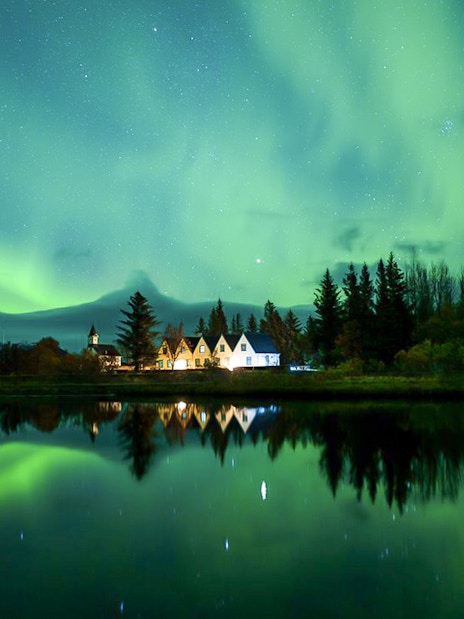 Northern lights over a serene landscape with trees and a small village, seen during a Super Jeep tour.