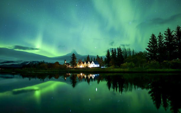 Northern lights over a serene landscape with trees and a small village, seen during a Super Jeep tour.