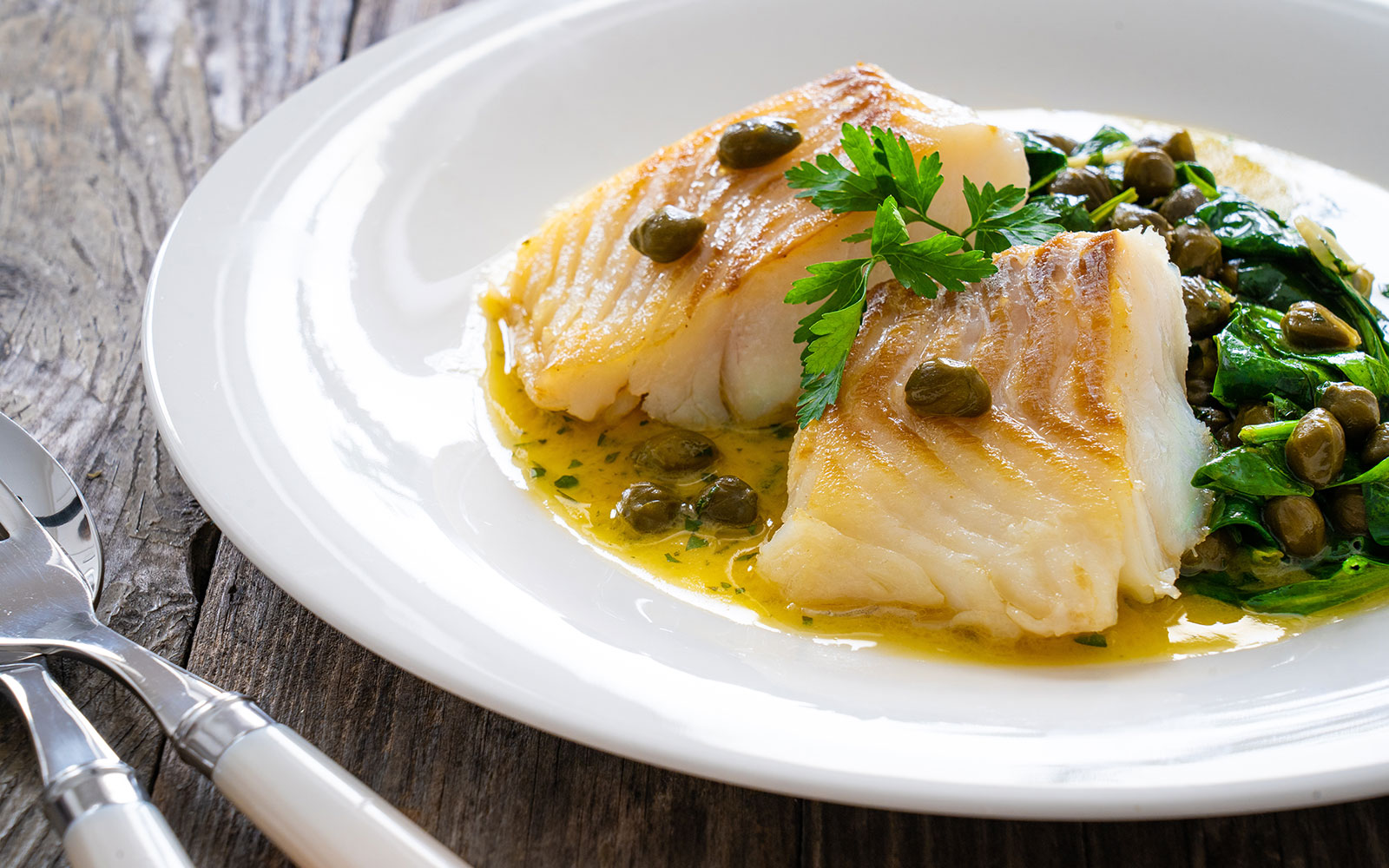 Fried cod with spinach and capers in saffron sauce on a wooden table.