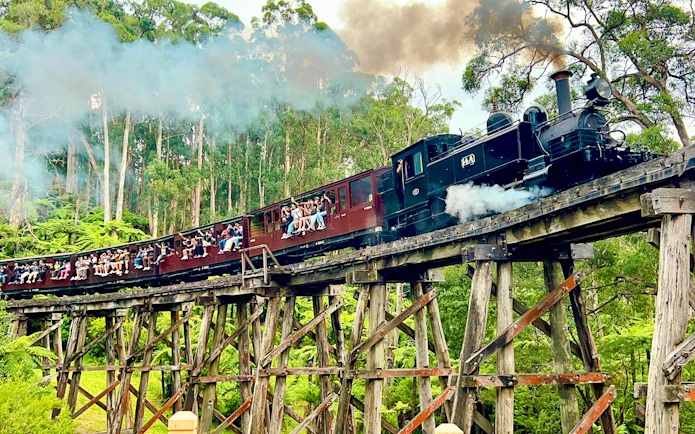 Puffing Billy steam train on a wooden trestle bridge with passengers sitting on carriage sills.