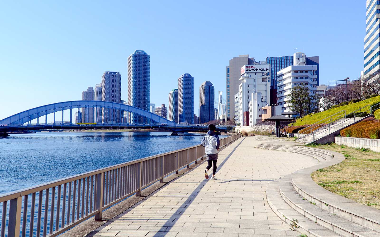 Jogger on Sumida riverside path with Tokyo skyline and bridge in view.