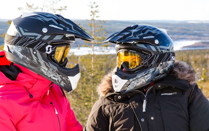Two people in winter gear with helmets, preparing for a snowmobile sprint in Rovaniemi.