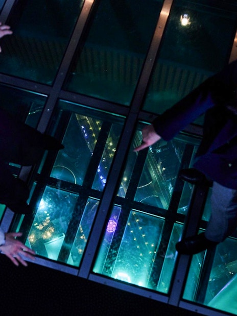 Visitors standing on glass floor at Tembo Deck, night view below.