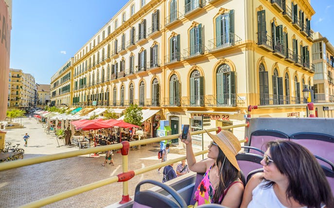 Passengers on Malaga hop-on hop-off tour bus passing historic buildings.