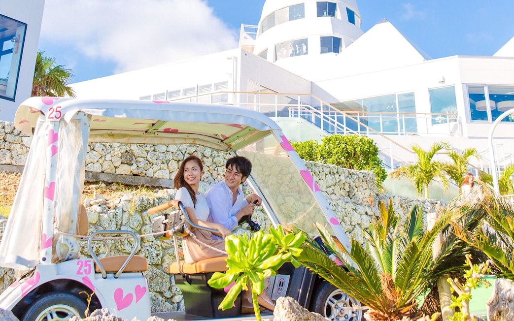 Couple driving decorated golf cart towards modern hotel at Hakone Kowakien Yunessun.