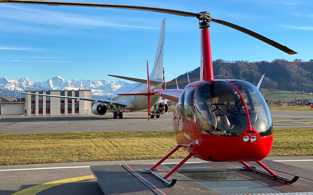 Red helicopter on tarmac at Bern-Belp Airport with Stockhorn Mountain in background.